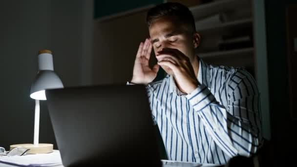 Уставший молодой человек с головной болью работает допоздна на ноутбуке в тускло освещенном офисе. - Кадры, видео