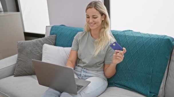 Блондинка с кредиткой и смехом во время работы над ноутбуком в современной гостиной - Кадры, видео