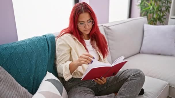 Kluge junge rothaarige Frau liest Literatur, vertieft in Wissen, sitzt gemütlich auf dem Sofa im heimischen Wohnzimmer und schafft eine entspannte und doch ernste Kulisse - Filmmaterial, Video
