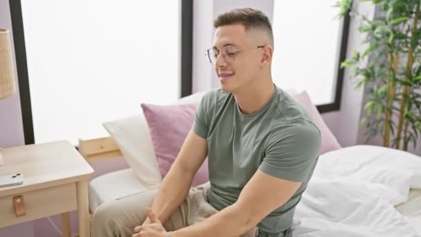Jeune homme hispanique souriant portant des lunettes assis avec désinvolture dans une chambre à coucher avec un intérieur confortable, respirant une aura détendue et attrayante. - Séquence, vidéo