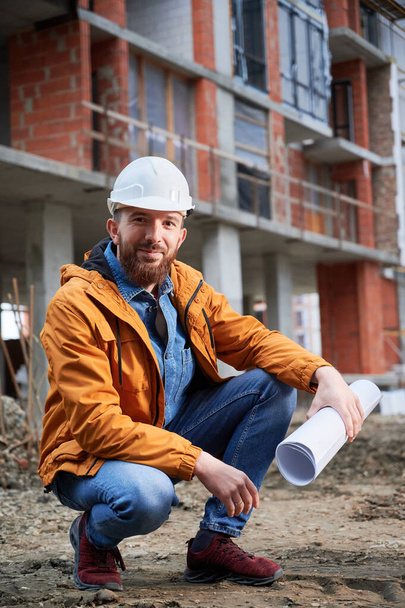 Fröhlicher Mann mit Schutzhelm, der einen Bauplan in der Hand hält und lächelt, während er auf der Baustelle hockt. Fröhliche männliche Bauarbeiter posieren vor Gebäude im Bau. - Foto, Bild