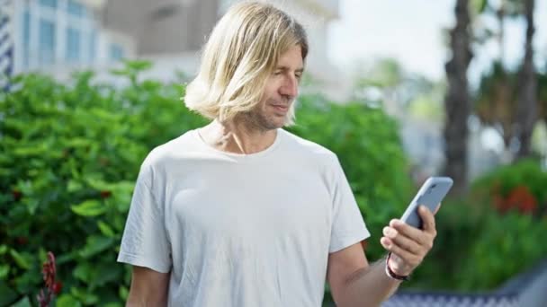 Красивий чоловік з довгим світлим волоссям використовує смартфон і святкує успіх на відкритому повітрі в пишному зеленому міському парку. - Кадри, відео