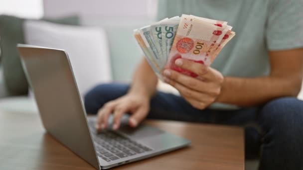 Joven, sentado en su sofá en casa, manejando billetes de yuan mientras está absorto en las finanzas en línea en su computadora portátil - Imágenes, Vídeo