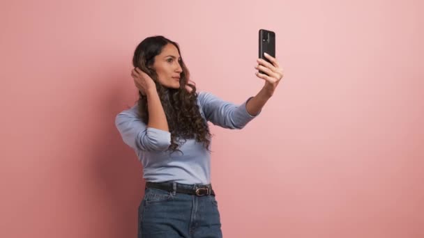Video met roze achtergrond van een volwassen Spaanse vrouw poseren en glimlachen tijdens het nemen van selfie met mobiele telefoon - Video