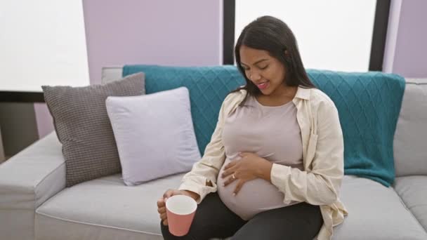 Świecąca młoda kobieta w ciąży, błogo uśmiechnięta, pije poranną kawę w domu, z miłością dotykając brzucha - Materiał filmowy, wideo