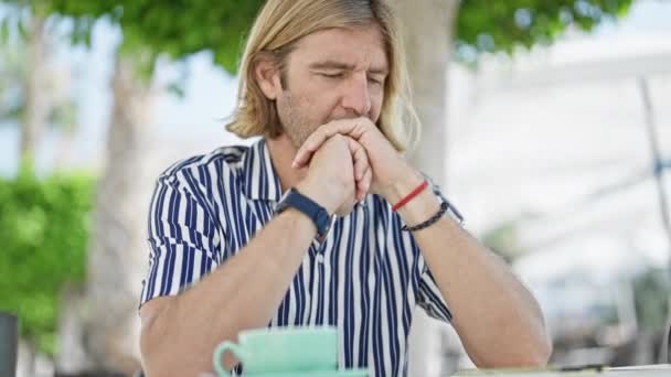 Thoughtful man with long hair sitting at an outdoor cafe, contemplating while surrounded by urban greenery. - Footage, Video