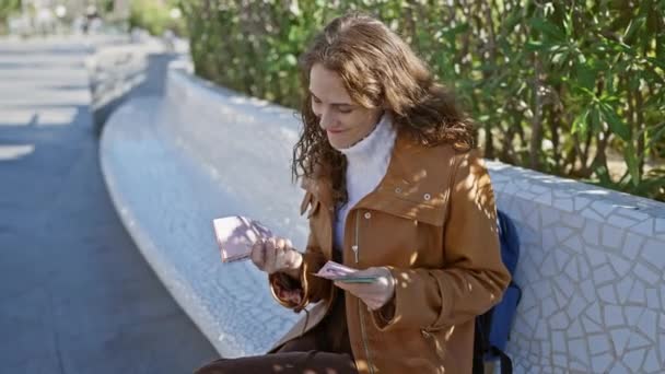 Μια χαρούμενη νεαρή γυναίκα εξετάζει αυτά τα χαρτονομίσματα ενώ κάθεται σε ένα παγκάκι σε εξωτερικό χώρο περιτριγυρισμένο από τη φύση. - Πλάνα, βίντεο