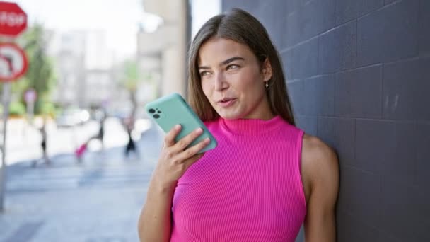 Atrakcyjna młoda Latynoska kobieta radośnie wysyła wiadomość dźwiękową przez smartfona, opierając się swobodnie o miejski mur uliczny, jej pogodny uśmiech oświetlający jej piękną twarz - Materiał filmowy, wideo