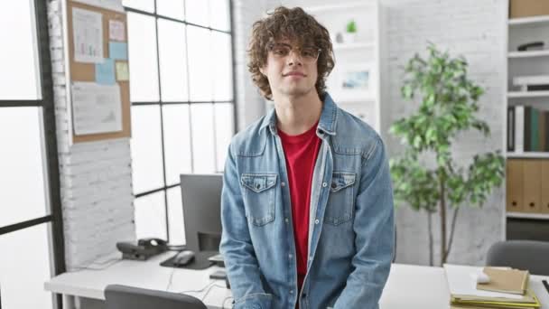 Komea nuori mies, jolla on kiharat hiukset ja lasit, seisoo luottavaisesti modernissa toimistossa. - Materiaali, video