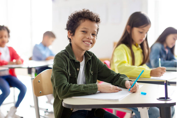 Vidám iskolás fiú ül az asztalánál, mosolyog a kamera előtt, miközben dolgozatot ír, körülvéve osztálytársakkal, akik koncentrált tanulmányokat folytatnak. - Fotó, kép