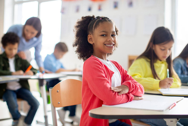 Felice studentessa nera si siede alla scrivania con le braccia incrociate, sorridendo alla fotocamera, ambiente scolastico vibrante e positivo - Foto, immagini