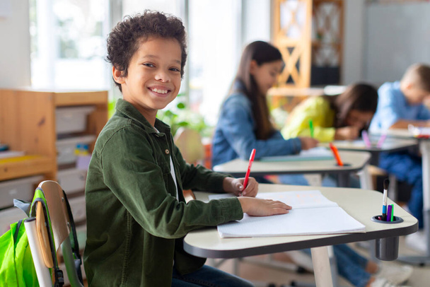Lächelnder schwarzer Junge, der im Klassenzimmer schreibt, lernt, lernt und lernt. Latein männlicher Schüler in der Schule, Vorbereitung auf Prüfung und Projekt - Foto, Bild