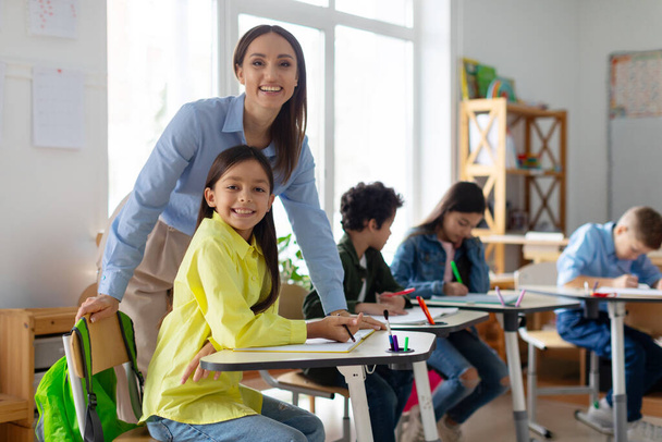 Fröhliche Lehrerin arbeitet mit Grundschulmädchen am Schreibtisch. Glückliche Lehrer und Mädchen lächeln in die Kamera im Klassenzimmer - Foto, Bild