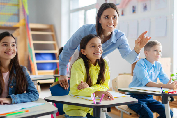 Vrolijke leraar staat tussen geëngageerde leerlingen, wijzend naar schoolbord terwijl het uitleggen, het creëren van een interactieve en levendige leersfeer - Foto, afbeelding
