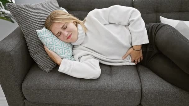 Donna bionda che sperimenta mal di stomaco mentre riposa su un divano grigio all'interno, esprimendo disagio. - Filmati, video
