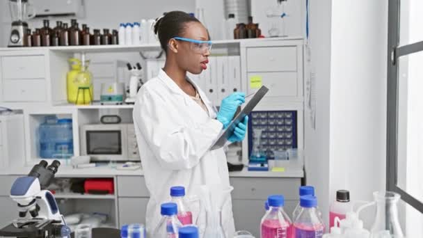 Appassionata scienziata afroamericana in guanti da laboratorio, affascinata dal potere della ricerca medica, profondamente assorta nel prendere appunti in un laboratorio di scienza indoor - Filmati, video