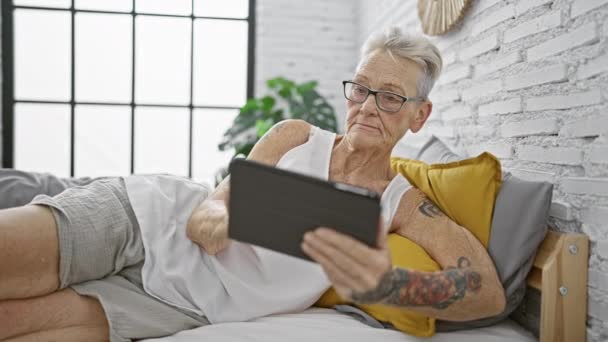 Femme âgée aux cheveux gris confiante couchée au lit, profitant d'une détente matinale avec pavé tactile dans une chambre confortable - Séquence, vidéo