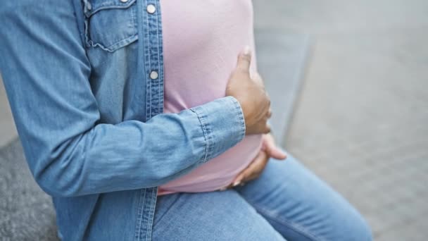 Emotionale junge schwangere Frau, berührt von der Mutterschaft, sitzt sorglos auf einer Straßenbank in der Stadt und wiegt ihren wachsenden Bauch im Sonnenlicht. - Filmmaterial, Video
