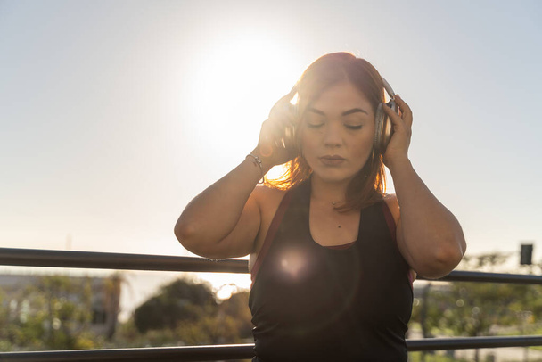 Споглядальна молода жінка насолоджується мирною миттю, слухаючи її навушники, як заходить сонце, оточуючи її теплим сяйвом. - Фото, зображення