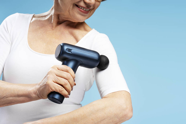 Atractiva mujer mayor sonriente, abuela moderna feliz usando pistola de masaje para los músculos, haciendo auto masaje aislado sobre fondo azul  - Foto, Imagen