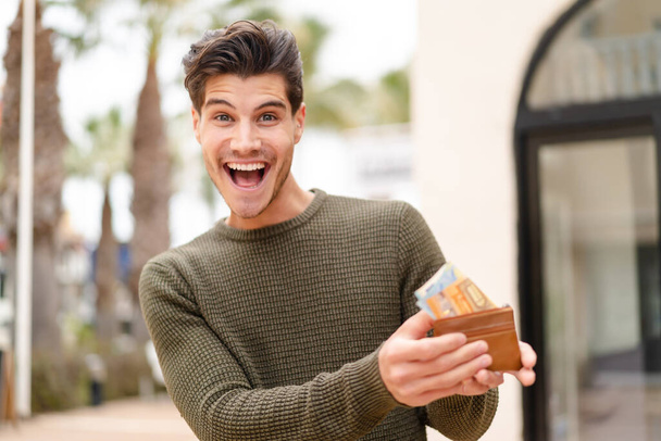 Fiatal fehér férfi a szabadban, pénztárcával a kezében, boldog arckifejezéssel. - Fotó, kép