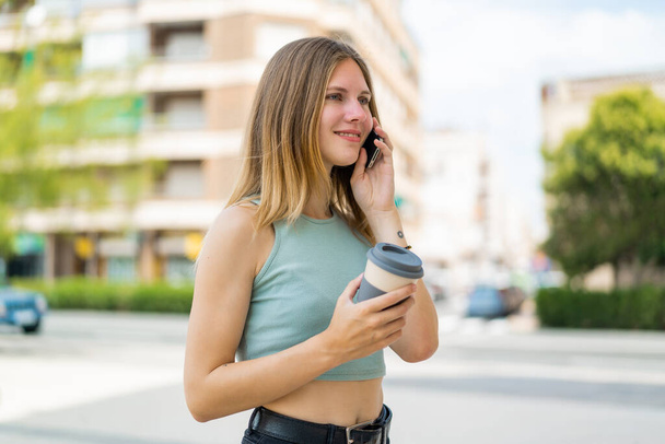 Giovane donna bionda all'aperto utilizzando il telefono cellulare e tenendo un caffè con espressione felice - Foto, immagini