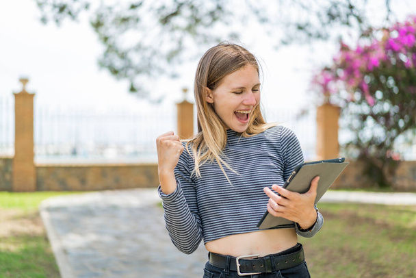Young blonde woman holding a tablet at outdoors celebrating a victory - Photo, Image