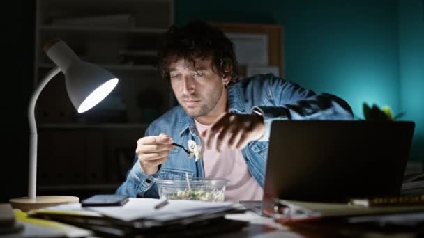 Keskittynyt mies moniajo hämärässä kotitoimistossa, syö salaattia työskennellessään myöhään yöllä. - Materiaali, video