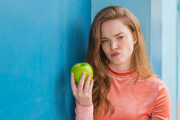 Νεαρή κοκκινομάλλα γυναίκα με ένα μήλο σε εξωτερικούς χώρους με θλιβερή έκφραση - Φωτογραφία, εικόνα