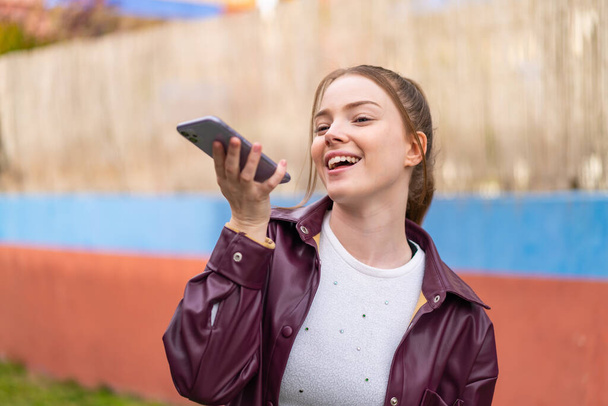 Νεαρή όμορφη κοπέλα χρησιμοποιώντας το κινητό τηλέφωνο σε εξωτερικούς χώρους κάνοντας χειρονομία αμφιβολίες, ενώ σηκώνοντας τους ώμους - Φωτογραφία, εικόνα