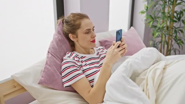 Μια χαλαρή γυναίκα με ριγέ πουκάμισο απολαμβάνει τη χρήση του smartphone της σε ένα άνετο υπνοδωμάτιο ρύθμιση. - Πλάνα, βίντεο