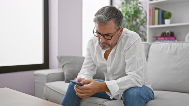Jeune homme hispanique aux cheveux gris utilisant un smartphone assis sur un canapé à la maison - Séquence, vidéo