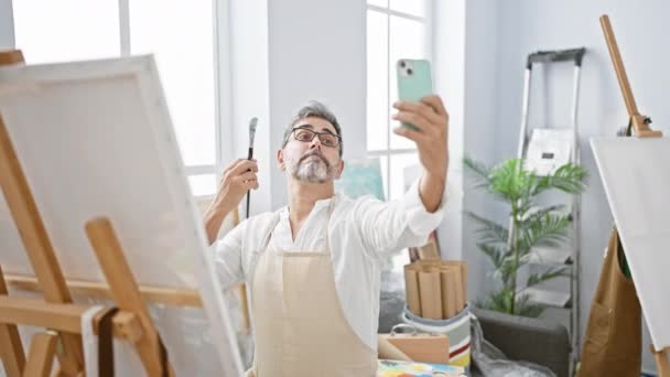 Sorrindo jovem, artista hispânico de cabelos grisalhos captura uma selfie alegre com smartphone em meio a pincéis e telas em estúdio de arte - Filmagem, Vídeo