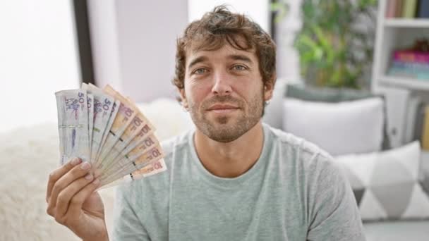 Joven alegre mostrando una señal de ok, sosteniendo un puñado de pesos colombianos en su casa fresca, exudando confianza y alegría - Imágenes, Vídeo