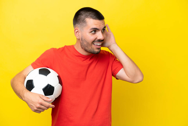 Młody biały mężczyzna gra w piłkę nożną odizolowany na żółtym tle słuchając czegoś kładąc rękę na uchu - Zdjęcie, obraz
