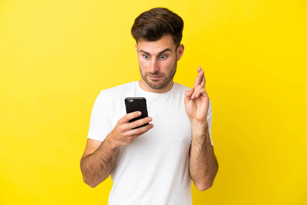 Jonge blanke knappe man geïsoleerd op gele achtergrond met behulp van mobiele telefoon met vingers kruisen - Foto, afbeelding