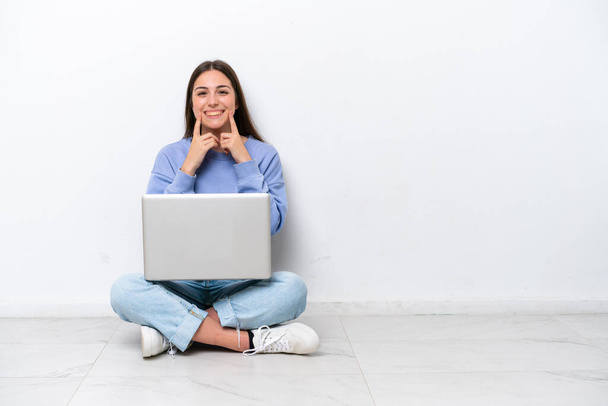 Giovane donna caucasica con computer portatile seduto sul pavimento isolato su sfondo bianco sorridente con un'espressione felice e piacevole - Foto, immagini