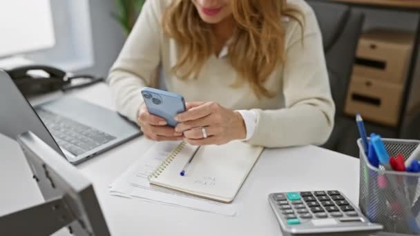 Mujer sonriente usando teléfono inteligente en un entorno de oficina moderno con computadora portátil y papelería alrededor. - Imágenes, Vídeo