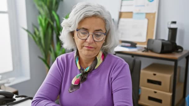 Una mujer madura comprueba la hora en un ambiente de oficina bien iluminado, con gafas y un suéter púrpura. - Imágenes, Vídeo