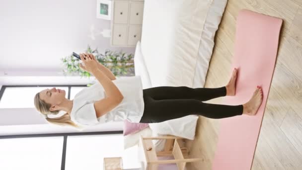 Молода жінка в активному одязі взаємодіє зі смартфоном у яскравій спальні з килимом для йоги - Кадри, відео