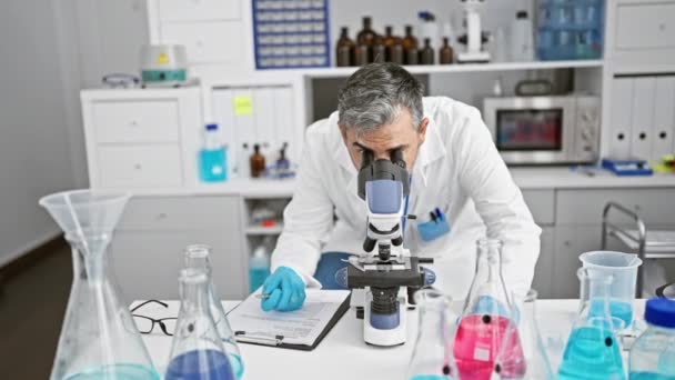 Passionate young hispanic, grey-haired man, an attractive scientist, engrossed in medical research at laboratory. vigorously taking notes, intensely peering through microscope in serious analysis. - Footage, Video