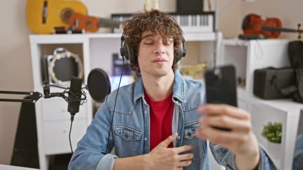 Wesoły człowiek w dżinsowej kurtce vlogs ze smartfonem w studiu muzycznym z gitarami i sprzętem do nagrywania. - Materiał filmowy, wideo