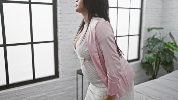 Nuori raskaana oleva nainen, joka seisoo sisätiloissa, heittää varovasti katseensa kohti makuuhuoneensa ikkunaa, kädet lepäävät vatsallaan. - Materiaali, video