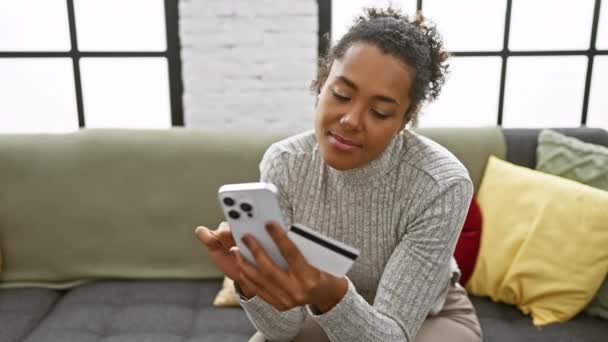 Uma mulher sorridente usando smartphone e segurando cartão de crédito em um sofá cinza com almofadas coloridas. - Filmagem, Vídeo
