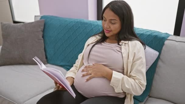 Hehkuva nuori raskaana oleva nainen nauttii mukavasta levosta, syventynyt lukemaan kirjaa koskettaessaan vatsaa ja säteilevää positiivisuutta kotona - Materiaali, video