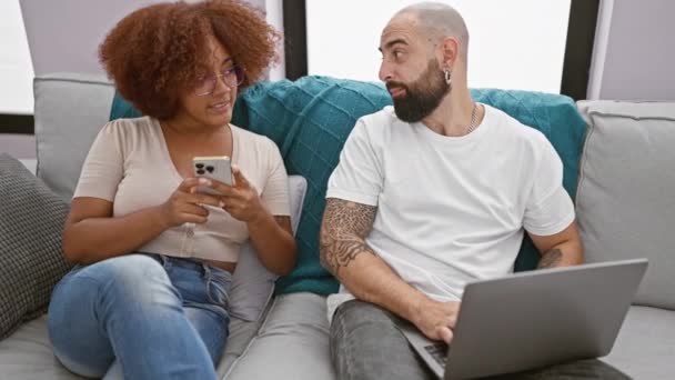 Όμορφο ζευγάρι κάθεται μαζί στο σαλόνι, χαμογελώντας και με αυτοπεποίθηση πληκτρολογώντας μηνύματα στο laptop και smartphone στο σπίτι - Πλάνα, βίντεο