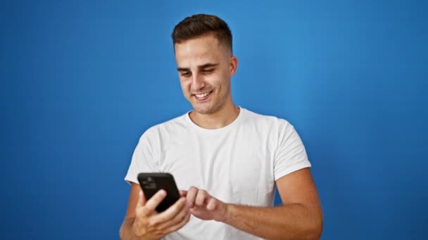Uśmiechnięty młody Hiszpan używający smartfona przeciwko odizolowanej niebieskiej ścianie - Materiał filmowy, wideo