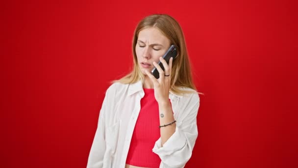 Una mujer rubia con ojos azules reacciona emocionalmente mientras habla en un teléfono inteligente sobre un fondo rojo vibrante. - Imágenes, Vídeo