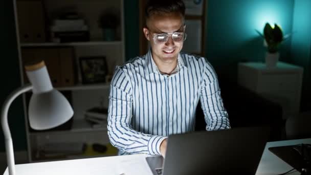 Профессиональный латиноамериканец работает допоздна за рабочим столом, сосредоточенный на ноутбуке в современном рабочем месте. - Кадры, видео