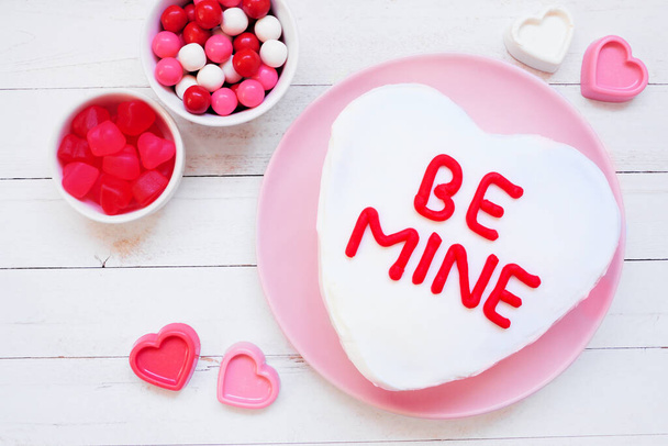 Fun Valentines Day BE MIJNE hartvormige taart met witte glazuur. Bovenaanzicht over een witte houten achtergrond. - Foto, afbeelding